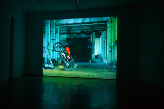 Video Invitational #3 - Video in tutti i sensi, Hubert Dobler, Bull, 2005

Un’azione che entra in loop, un’immagine tutt’altro che statica in cui una moto da cross viene bloccata attraverso tiranti elastici al soffitto di uno spazio industriale dismesso. Appare un cavallo imbizzarrito che vuole faticosamente liberarsi dalla propria prigionia. Tanto più violente sono le accellerate che si autogenerano e i conseguenti tentativi di fuga, tanto più fallimentari le inesorabili ricadute sui propri passi. È di scena il faticoso tentativo di uscire dalla propria pelle.