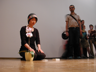 As soon as possible. Performance loop. The Class of Marina Abramovic - Braunschweig School of Art, Heejung Um, "Love Odyssey".

Artist kneels(on his knees).SHE Takes a white paper and puts it down at the middle of the distance to the orchid flowerpot. She takes a small piece of bark from the flowerpot and places it on the white paper.he takes another white paper and places it on top of the piece of bark (sandwich). This action is repeated. She builds the tower with papers and pieces of bark from the orchid flowerpot.